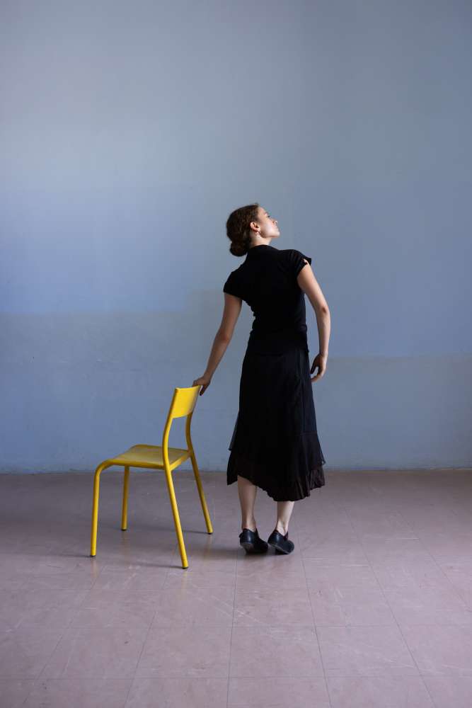 Photo d'une jeune-femme sur fond bleu tenant une chaise jaune avec une attitude de danseuse